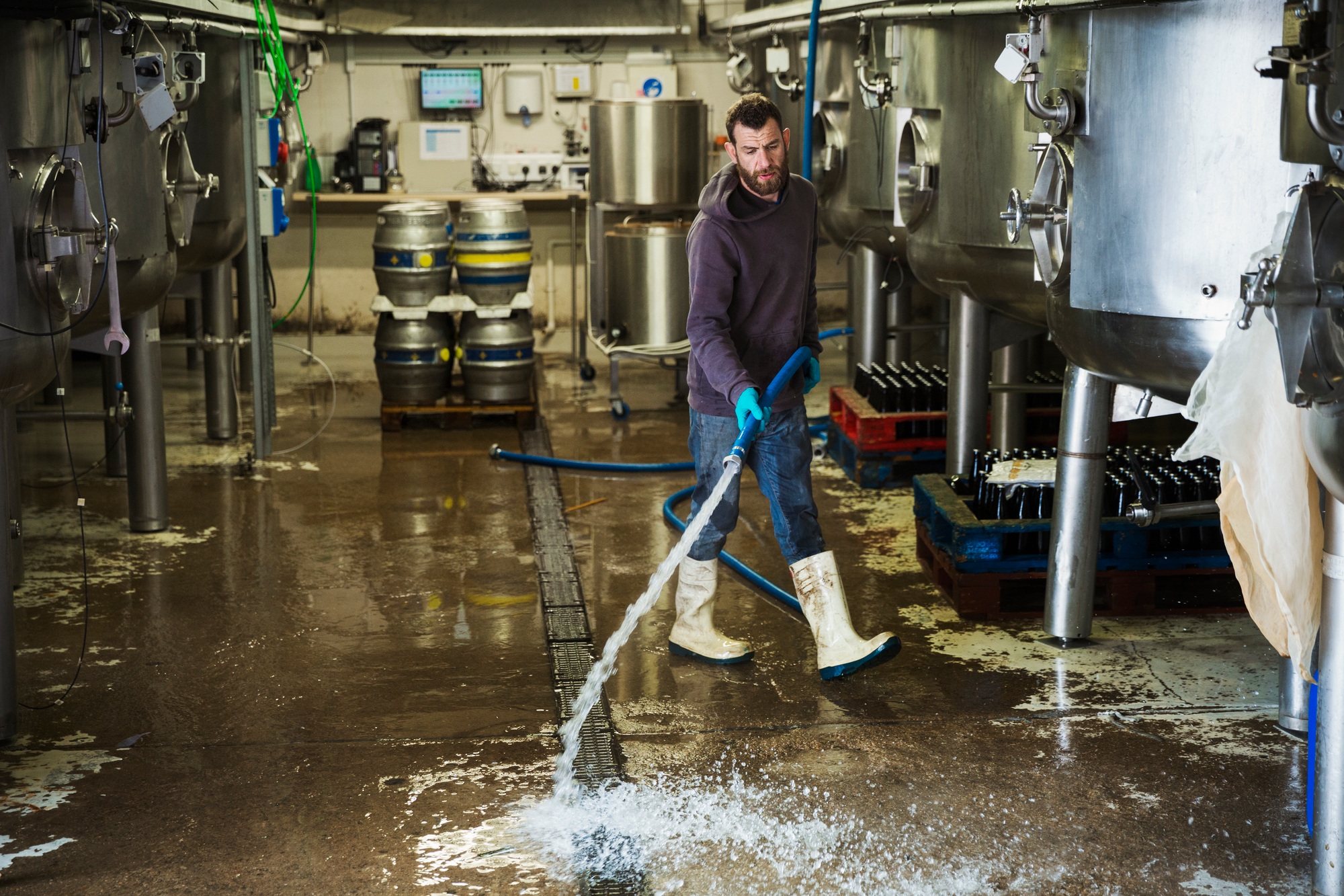 Man working in a brewery, cleaning floor with water hose.